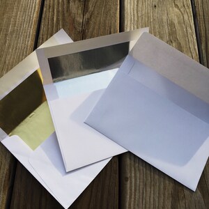 May include: Three white paper envelopes with open flaps. One envelope features a reflective gold foil lining, another displays a silver foil lining, and the third envelope is plain white on the inside. The envelopes are arranged in an overlapping stack.