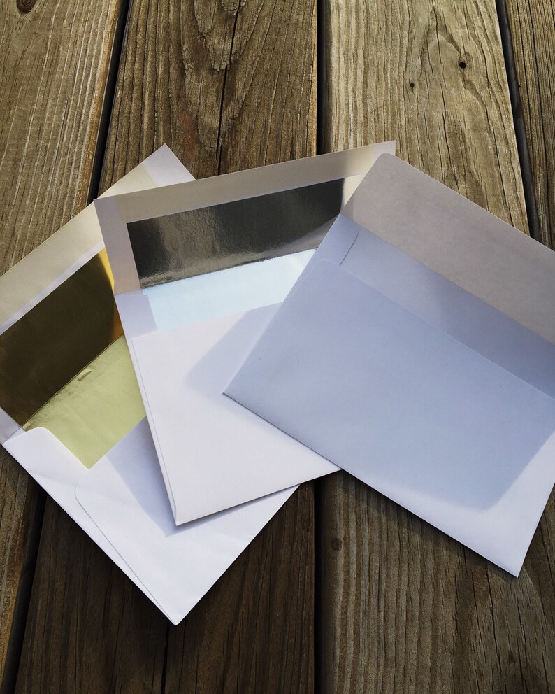 May include: Three white paper envelopes with open flaps. One envelope features a reflective gold foil lining, another displays a silver foil lining, and the third envelope is plain white on the inside. The envelopes are arranged in an overlapping stack.