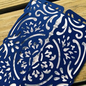 May include: A dark blue laser-cut paper invitation sleeve featuring an intricate floral and scrollwork pattern. The sleeve is folded over a white card insert with visible black printed text, showcasing a detailed die-cut design throughout.