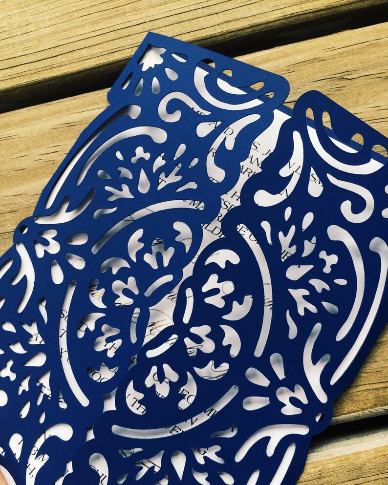 May include: A dark blue laser-cut paper invitation sleeve featuring an intricate floral and scrollwork pattern. The sleeve is folded over a white card insert with visible black printed text, showcasing a detailed die-cut design throughout.