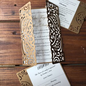 May include: A gold laser-cut gatefold wedding invitation set with white inserts featuring black text. The invitation includes details for Rita Fayne and Max Dovg, a reception card, and an RSVP card with lines for guest names and attendance status.
