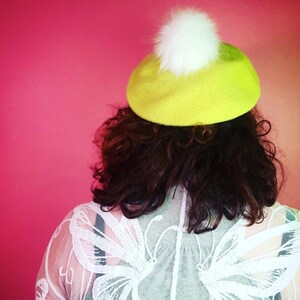 May include: A woman wearing a green beret with a white pom-pom and a white lace top with butterfly embroidery.