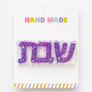May include: A purple glittery hair clip with the Hebrew word "Shabbat" on a white card. The card has the words "HAND MADE" in colorful letters at the top and a striped design at the bottom.