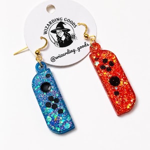 May include: A pair of blue and red glittery earrings shaped like game controllers. The blue earring has black buttons and the red earring has black buttons. The earrings are attached to gold-colored hooks. The earrings are hanging from a white tag with the text "WIZARDING GOODS" and the username "@wizarding_goods".