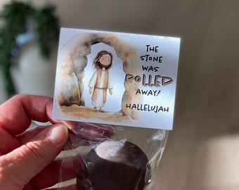 Può includere: Un piccolo uovo di cioccolato marrone avvolto in plastica trasparente con un'etichetta che recita "La pietra è stata rotolata via, Alleluia!" e presenta un'illustrazione a cartoni animati di Gesù che emerge da una grotta.