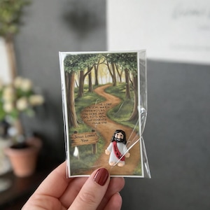 May include: A small, clear plastic package containing a card with a forest scene and a miniature Jesus figurine. The card has text that reads "The Lord Keeps Watch Over You As You Come and Go, Both Now and Forever. Psalm 1218" and "Jesus Loves You."
