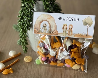 Può includere: Un sacchetto di plastica trasparente pieno di una varietà di snack, tra cui pretzel, cracker a forma di pesce rosso e caramelle. Un'etichetta bianca con le parole "HE is RISEN" è attaccata al sacchetto. L'etichetta presenta un'illustrazione ad acquerello di un arco di pietra, due figure e un albero.