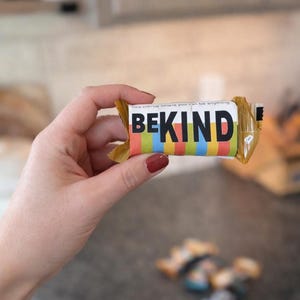 May include: A wrapped bar with the words "BE KIND" in bold black letters. The bar has a colorful striped design in red, orange, yellow, green, blue, and black. The packaging also includes the phrase "free clinic where you can be anything".