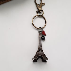May include: A bronze Eiffel Tower keychain with a red heart and black bead charm attached to a bronze key ring and clasp.