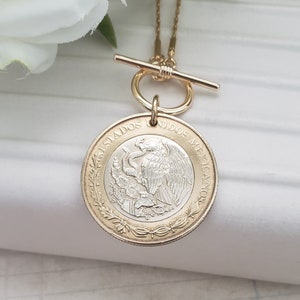 K&ouml;nnte beinhalten: Eine goldfarbene Knebelkette mit einem runden Anh&auml;nger aus einer mexikanischen M&uuml;nze. Die M&uuml;nze zeigt den Text ESTADOS UNIDOS MEXICANOS und einen Adler auf einem Kaktus mit einer Schlange im Schnabel auf silberfarbenem Grund.
