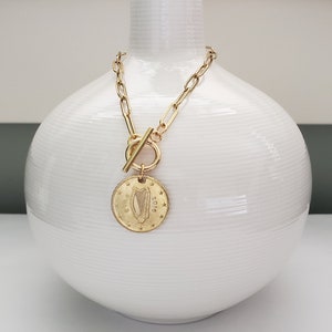 May include: A gold chain necklace with a toggle clasp and a round coin pendant featuring a harp design and the year 2012. The necklace is displayed on a white ceramic vase.
