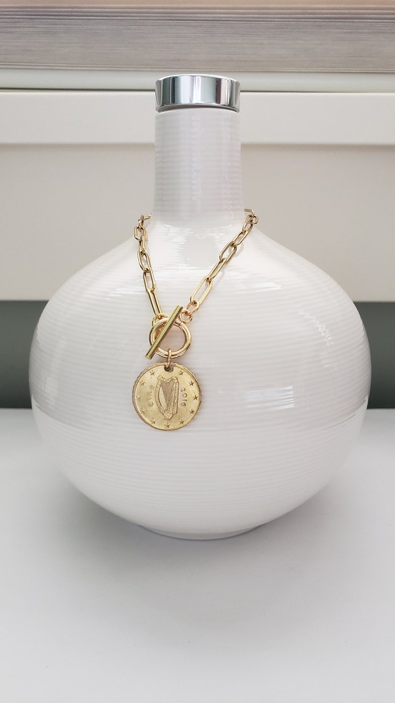 May include: A gold chain necklace with a toggle clasp and a round coin pendant featuring a harp design and the year 2012. The necklace is displayed on a white ceramic vase.