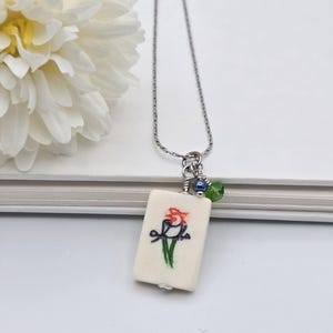 May include: A silver-toned chain necklace featuring a rectangular off-white pendant with a hand-painted bird illustration in red, black, and green. The pendant is accented with a small dark blue bead and a faceted green crystal bead near the top.
