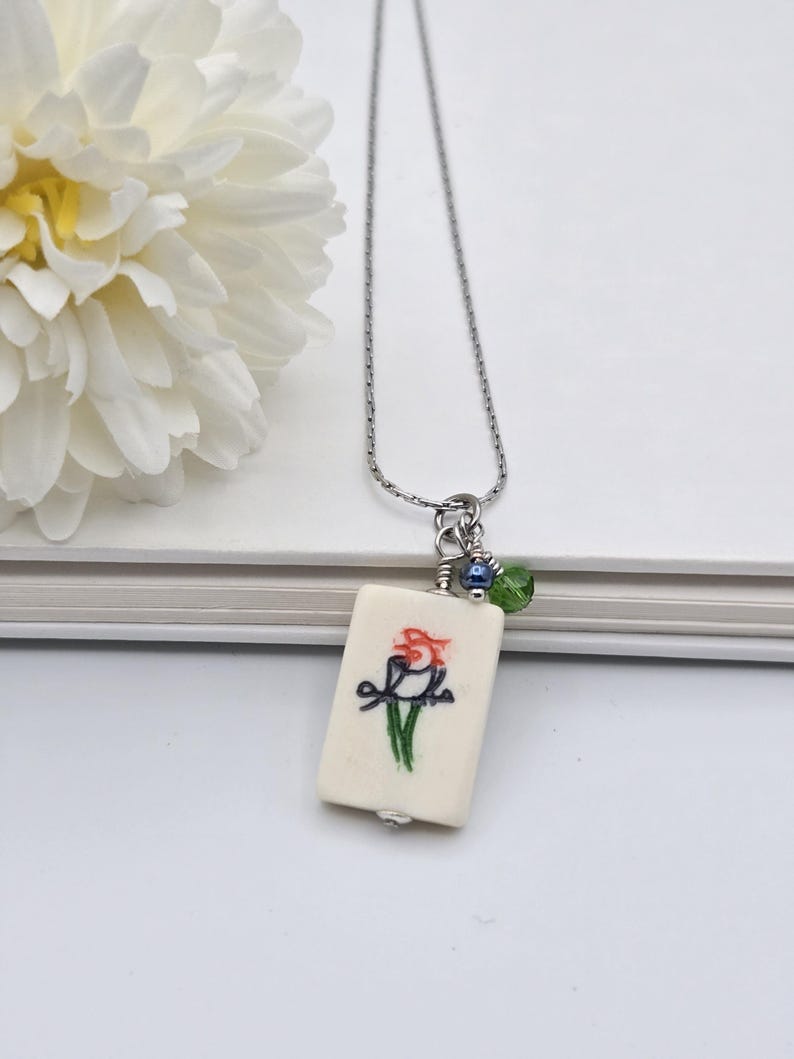 May include: A silver-toned chain necklace featuring a rectangular off-white pendant with a hand-painted bird illustration in red, black, and green. The pendant is accented with a small dark blue bead and a faceted green crystal bead near the top.