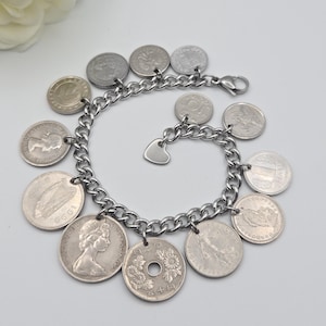 K&ouml;nnte beinhalten: Silberfarbenes Charm-Armband mit einer Kette, verziert mit verschiedenen Vintage-M&uuml;nzen. Das Armband hat einen kleinen Herzanh&auml;nger. Die M&uuml;nzen, in verschiedenen Gr&ouml;&szlig;en und Designs, verleihen dem Armband ein einzigartiges, antikes Aussehen.
