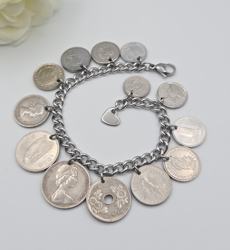 K&ouml;nnte beinhalten: Silberfarbenes Charm-Armband mit einer Kette, verziert mit verschiedenen Vintage-M&uuml;nzen. Das Armband hat einen kleinen Herzanh&auml;nger. Die M&uuml;nzen, in verschiedenen Gr&ouml;&szlig;en und Designs, verleihen dem Armband ein einzigartiges, antikes Aussehen.