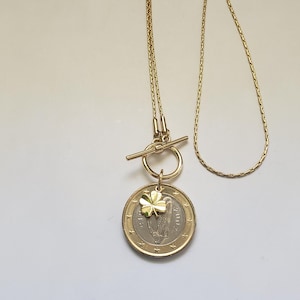 May include: A gold chain necklace with a toggle clasp and a pendant featuring a gold shamrock charm and a silver Irish euro coin with the harp of Ireland design.