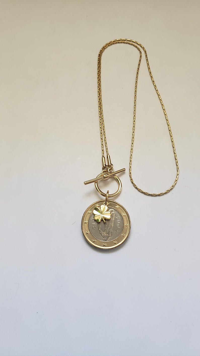 May include: A gold chain necklace with a toggle clasp and a pendant featuring a gold shamrock charm and a silver Irish euro coin with the harp of Ireland design.