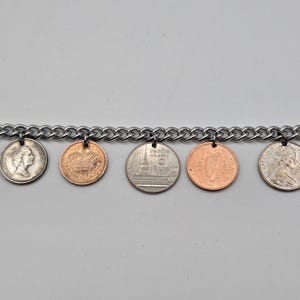 May include: A silver-toned metal chain bracelet featuring seven dangling coins of various sizes and colors, including copper-colored and silver-colored pieces. The coins are attached to the chain links with small metal jump rings.