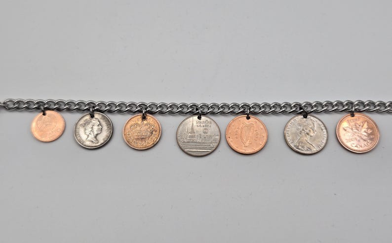 May include: A silver-toned metal chain bracelet featuring seven dangling coins of various sizes and colors, including copper-colored and silver-colored pieces. The coins are attached to the chain links with small metal jump rings.