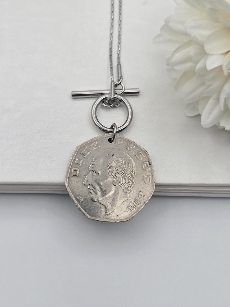 K&ouml;nnte beinhalten: Eine silberfarbene Halskette mit Knebelverschluss und einem Anh&auml;nger aus einer siebeneckigen mexikanischen Zehn-Pesos-M&uuml;nze. Die M&uuml;nze zeigt ein Portr&auml;t im Profil, den Schriftzug DIEZ PESOS und das Jahr 1982 an einer d&uuml;nnen Metallkette.
