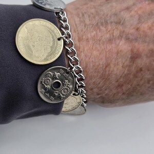 Pode incluir: Uma pulseira de corrente de metal prateado com v&aacute;rias moedas pendentes de v&aacute;rios tamanhos e cores, incluindo uma moeda dourada da Argentina e uma moeda japonesa prateada com um orif&iacute;cio central e grava&ccedil;&otilde;es florais.