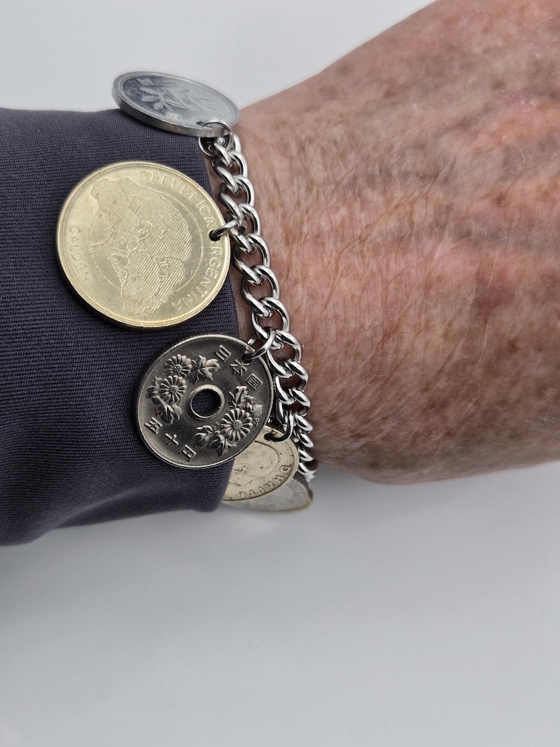 Pode incluir: Uma pulseira de corrente de metal prateado com v&aacute;rias moedas pendentes de v&aacute;rios tamanhos e cores, incluindo uma moeda dourada da Argentina e uma moeda japonesa prateada com um orif&iacute;cio central e grava&ccedil;&otilde;es florais.