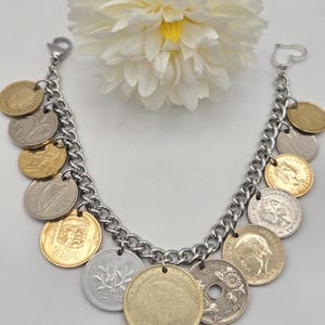 Pode incluir: Uma pulseira de corrente de metal prateado com v&aacute;rias moedas internacionais em tons de dourado e prateado. A pulseira inclui um fecho em forma de cora&ccedil;&atilde;o numa extremidade e um fecho lagosta, com moedas que exibem diversos retratos, s&iacute;mbolos e textos.