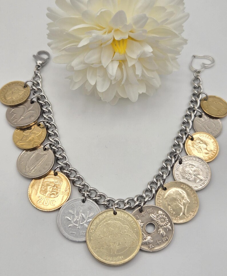 Pode incluir: Uma pulseira de corrente de metal prateado com v&aacute;rias moedas internacionais em tons de dourado e prateado. A pulseira inclui um fecho em forma de cora&ccedil;&atilde;o numa extremidade e um fecho lagosta, com moedas que exibem diversos retratos, s&iacute;mbolos e textos.