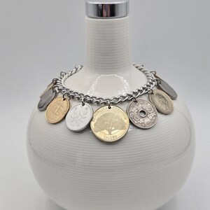 Pode incluir: Uma pulseira de corrente de metal prateado com v&aacute;rias moedas internacionais como pingentes, incluindo uma moeda dourada com um design de &aacute;rvore com a inscri&ccedil;&atilde;o Calden e uma moeda japonesa prateada com um motivo floral e o texto 50 ienes.
