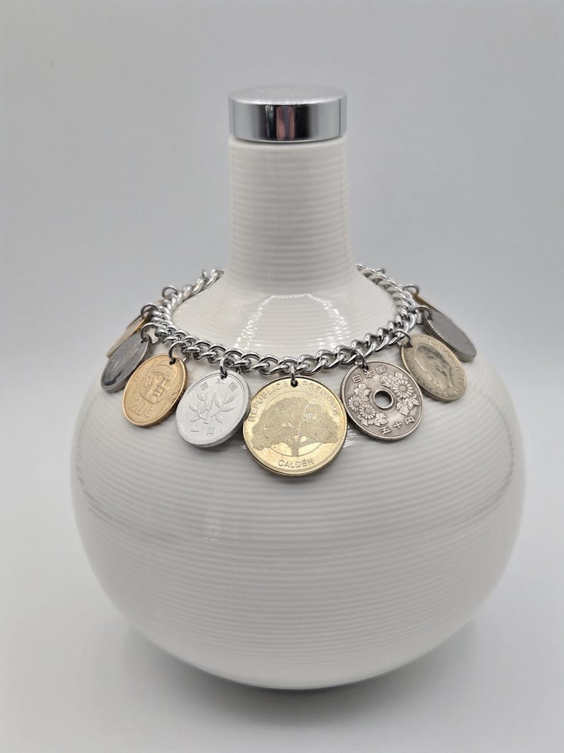 Pode incluir: Uma pulseira de corrente de metal prateado com v&aacute;rias moedas internacionais como pingentes, incluindo uma moeda dourada com um design de &aacute;rvore com a inscri&ccedil;&atilde;o Calden e uma moeda japonesa prateada com um motivo floral e o texto 50 ienes.