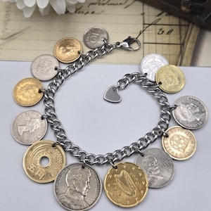 Pode incluir: Uma pulseira de elos de metal prateado com v&aacute;rias moedas internacionais como berloques. As moedas incluem pe&ccedil;as em tons de dourado e prateado com diversos retratos, s&iacute;mbolos e textos, finalizada com um pequeno berloque de cora&ccedil;&atilde;o prateado.