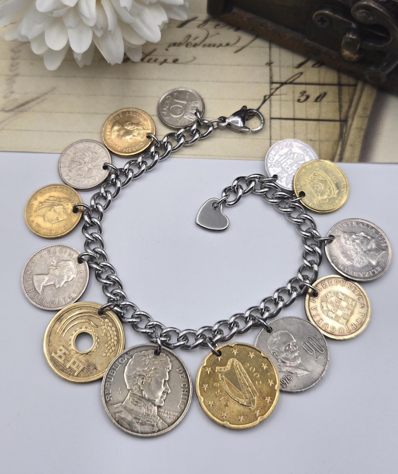 Pode incluir: Uma pulseira de elos de metal prateado com v&aacute;rias moedas internacionais como berloques. As moedas incluem pe&ccedil;as em tons de dourado e prateado com diversos retratos, s&iacute;mbolos e textos, finalizada com um pequeno berloque de cora&ccedil;&atilde;o prateado.