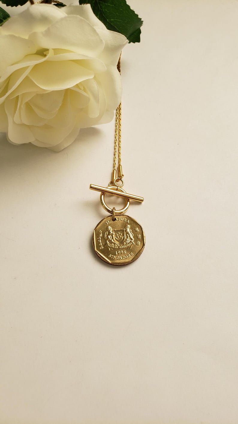May include: Gold necklace with a toggle clasp and a Singapore dollar coin pendant. The coin features the Singapore coat of arms and the year 1995.