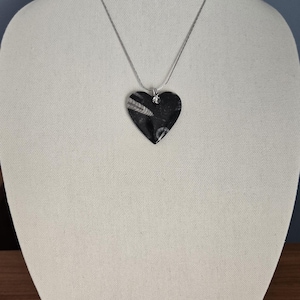 Könnte beinhalten: Eine herzförmige Anhängerkette aus einem schwarzen Stein mit sichtbaren weißen fossilen Muschelmustern. Der Anhänger hängt an einer dünnen, silberfarbenen Metallkette, die mit einem kleinen, klaren Kristallakzent am Herz befestigt ist.