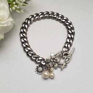 May include: A silver chain bracelet with a toggle clasp and charms, including a flower, a sunflower, and two pearl beads.