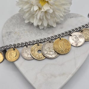 Pode incluir: Uma pulseira de corrente de metal prateado com uma cole&ccedil;&atilde;o de v&aacute;rias moedas internacionais como berloques. As moedas incluem pe&ccedil;as douradas e prateadas com diversos retratos, s&iacute;mbolos e textos, incluindo uma moeda japonesa de cinco ienes.