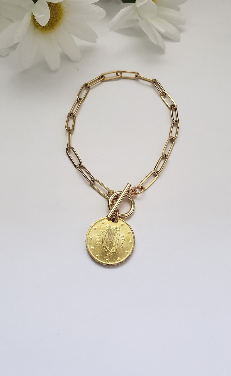 May include: Gold chain bracelet with a gold coin charm featuring the Irish harp and the year 2016. The coin is attached to the chain with a gold toggle clasp.