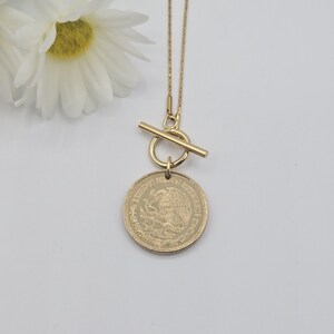K&ouml;nnte beinhalten: Eine goldfarbene Halskette mit einer d&uuml;nnen Kette und einem Knebelverschluss. Der Anh&auml;nger ist eine runde M&uuml;nze mit der Aufschrift ESTADOS UNIDOS MEXICANOS und einem gepr&auml;gten Adleremblem. Die Kette liegt neben wei&szlig;en Blumen.