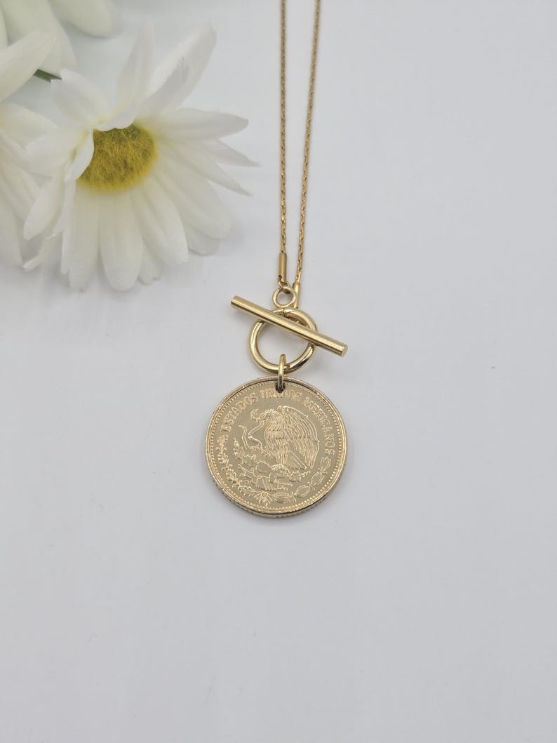 K&ouml;nnte beinhalten: Eine goldfarbene Halskette mit einer d&uuml;nnen Kette und einem Knebelverschluss. Der Anh&auml;nger ist eine runde M&uuml;nze mit der Aufschrift ESTADOS UNIDOS MEXICANOS und einem gepr&auml;gten Adleremblem. Die Kette liegt neben wei&szlig;en Blumen.