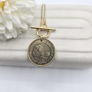 K&ouml;nnte beinhalten: Eine goldfarbene Halskette mit Knebelverschluss und rundem Anh&auml;nger. Der Anh&auml;nger ist eine M&uuml;nze mit einem auf einem Kaktus sitzenden Adler und der Inschrift ESTADOS UNIDOS MEXICANOS, umgeben von einem dekorativen Blattrand.