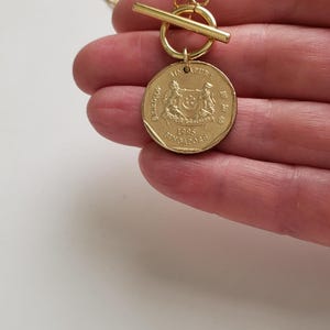 May include: Gold necklace with a toggle clasp and a Singapore dollar coin pendant. The coin features the Singapore coat of arms and the year 1995.
