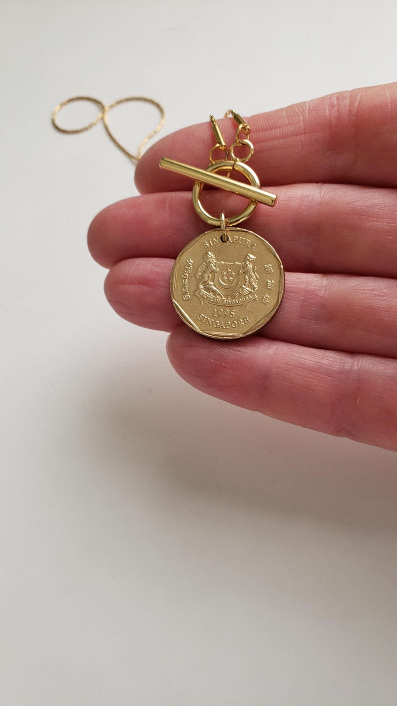 May include: Gold necklace with a toggle clasp and a Singapore dollar coin pendant. The coin features the Singapore coat of arms and the year 1995.