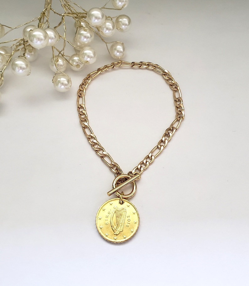 May include: A gold chain bracelet with a toggle clasp and a gold coin charm featuring the Irish harp and the year 2018.