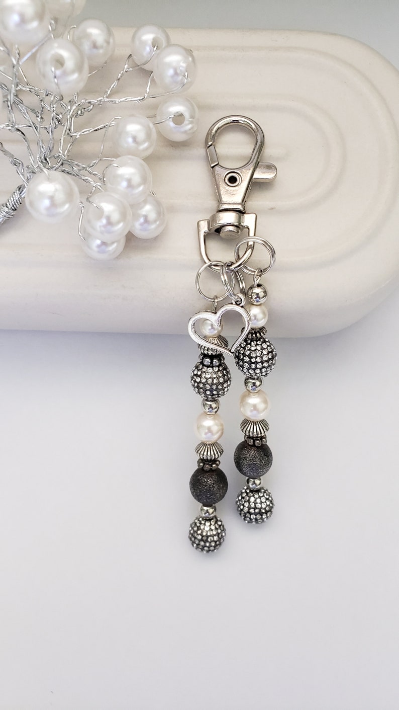 May include: A silver-toned keychain with two beaded charms. Each charm features a heart-shaped accent, pearl beads, and textured dark gray beads. The keychain also has a silver clasp.