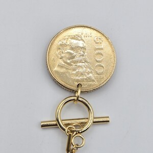 K&ouml;nnte beinhalten: Ein goldfarbener M&uuml;nzanh&auml;nger mit einem Portr&auml;t von Venustiano Carranza, dem Text $100 und dem Jahr 1983. Die M&uuml;nze ist an einem goldfarbenen Knebelverschluss mit einem kreisf&ouml;rmigen Ring und einem horizontalen Stab befestigt, der mit einer d&uuml;nnen Kette verbunden ist.