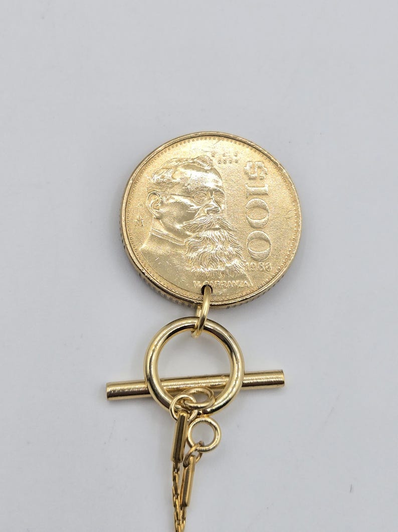 K&ouml;nnte beinhalten: Ein goldfarbener M&uuml;nzanh&auml;nger mit einem Portr&auml;t von Venustiano Carranza, dem Text $100 und dem Jahr 1983. Die M&uuml;nze ist an einem goldfarbenen Knebelverschluss mit einem kreisf&ouml;rmigen Ring und einem horizontalen Stab befestigt, der mit einer d&uuml;nnen Kette verbunden ist.