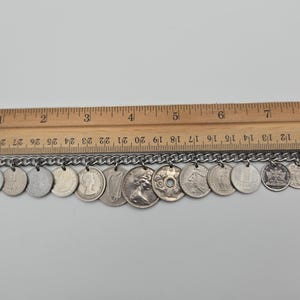 K&ouml;nnte beinhalten: Ein silbernes Armband mit einer Kette und verschiedenen M&uuml;nzanh&auml;ngern. Die M&uuml;nzen haben unterschiedliche Designs und Gr&ouml;&szlig;en. Ein Holzlineal liegt dar&uuml;ber und zeigt Messungen in Zentimetern an.