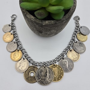 Pode incluir: Uma pulseira de berloques com uma corrente de metal prateado adornada com v&aacute;rias moedas internacionais em tons de dourado e prateado. As moedas exibem diversos retratos, s&iacute;mbolos e texto, incluindo uma moeda japonesa de cinco ienes com um furo.