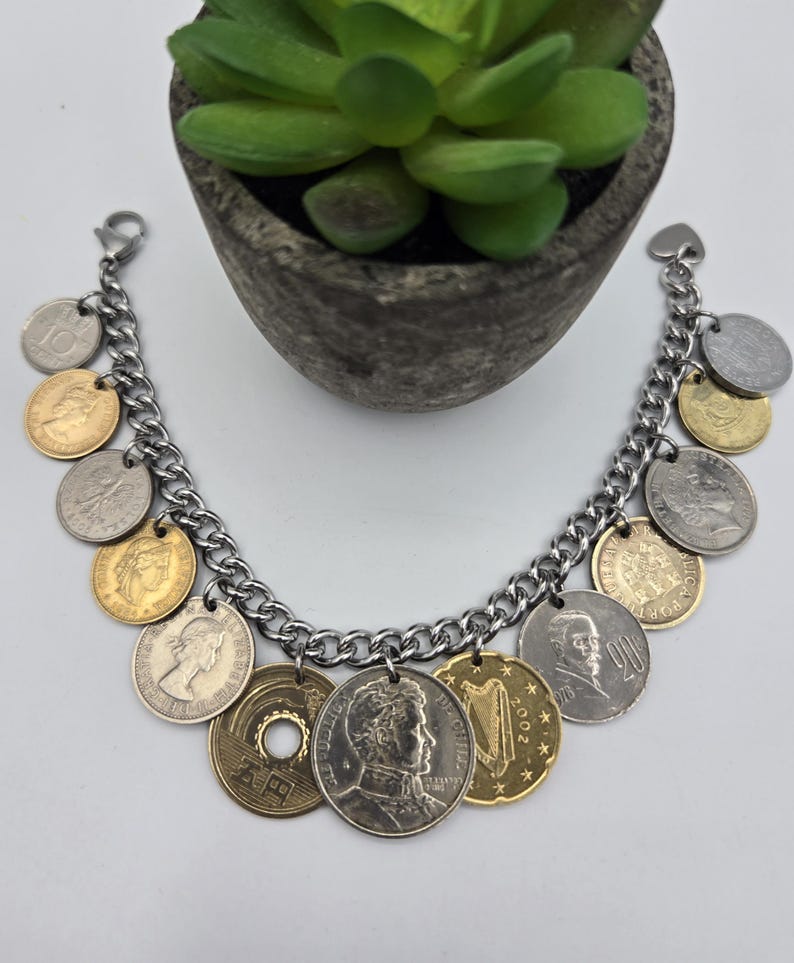 Pode incluir: Uma pulseira de berloques com uma corrente de metal prateado adornada com v&aacute;rias moedas internacionais em tons de dourado e prateado. As moedas exibem diversos retratos, s&iacute;mbolos e texto, incluindo uma moeda japonesa de cinco ienes com um furo.